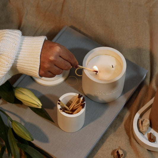 A person lighting a lavender scented candle with a match, surrounded by a cozy, relaxed setting with concrete jars and herbs.