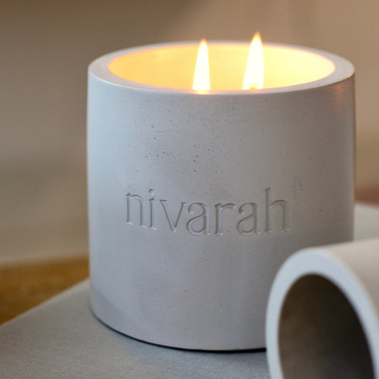 A grey candle with two wicks burning, encased in a concrete jar with the brand name 'nivarah' visible on the side.