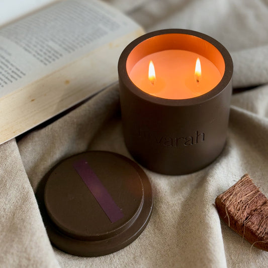 A mahogany teakwood scented candle with two wicks burning, placed inside a concrete jar on a textile surface, with an open lid beside it and a book in the background.