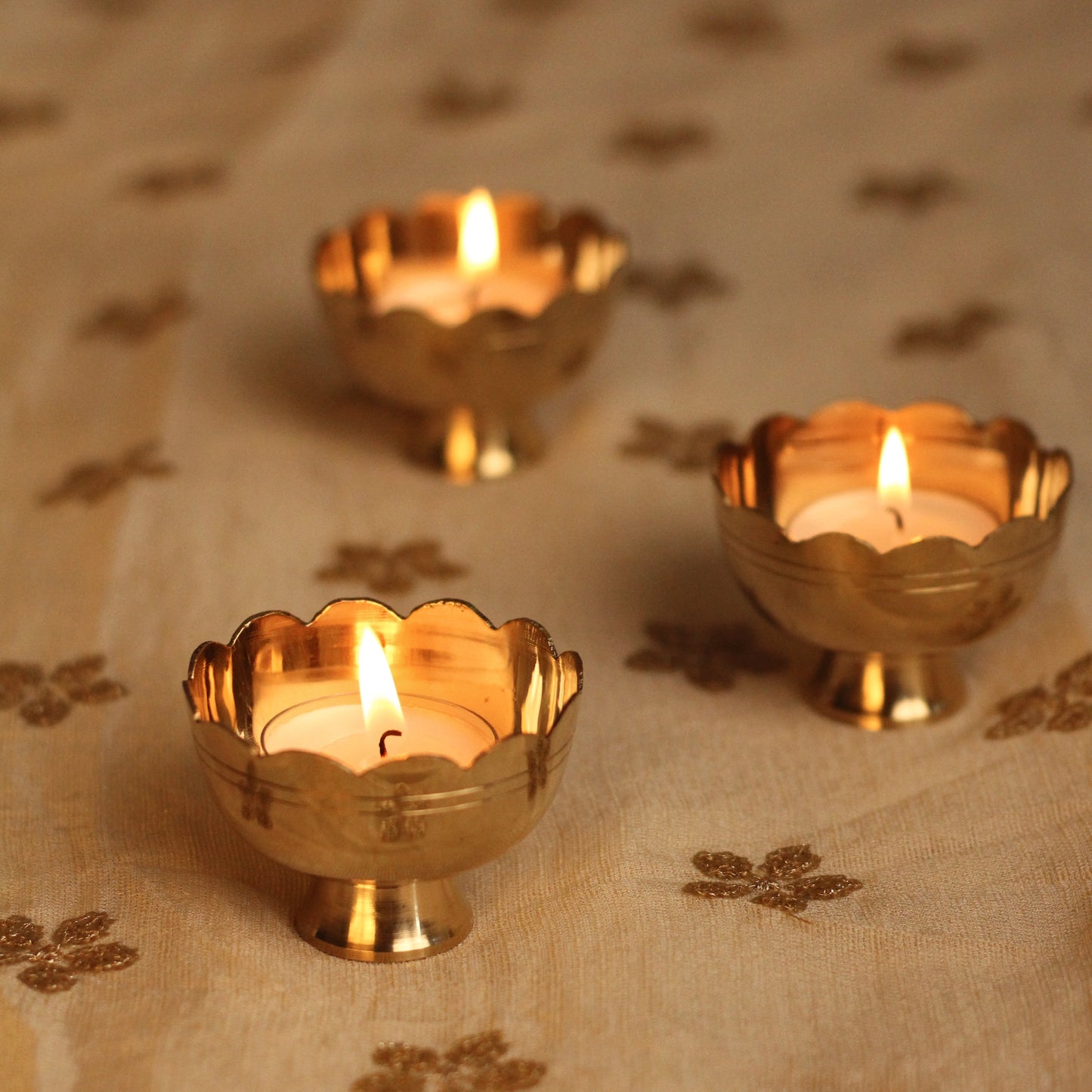 Three brass tealight holders with lit candles on a patterned fabric background