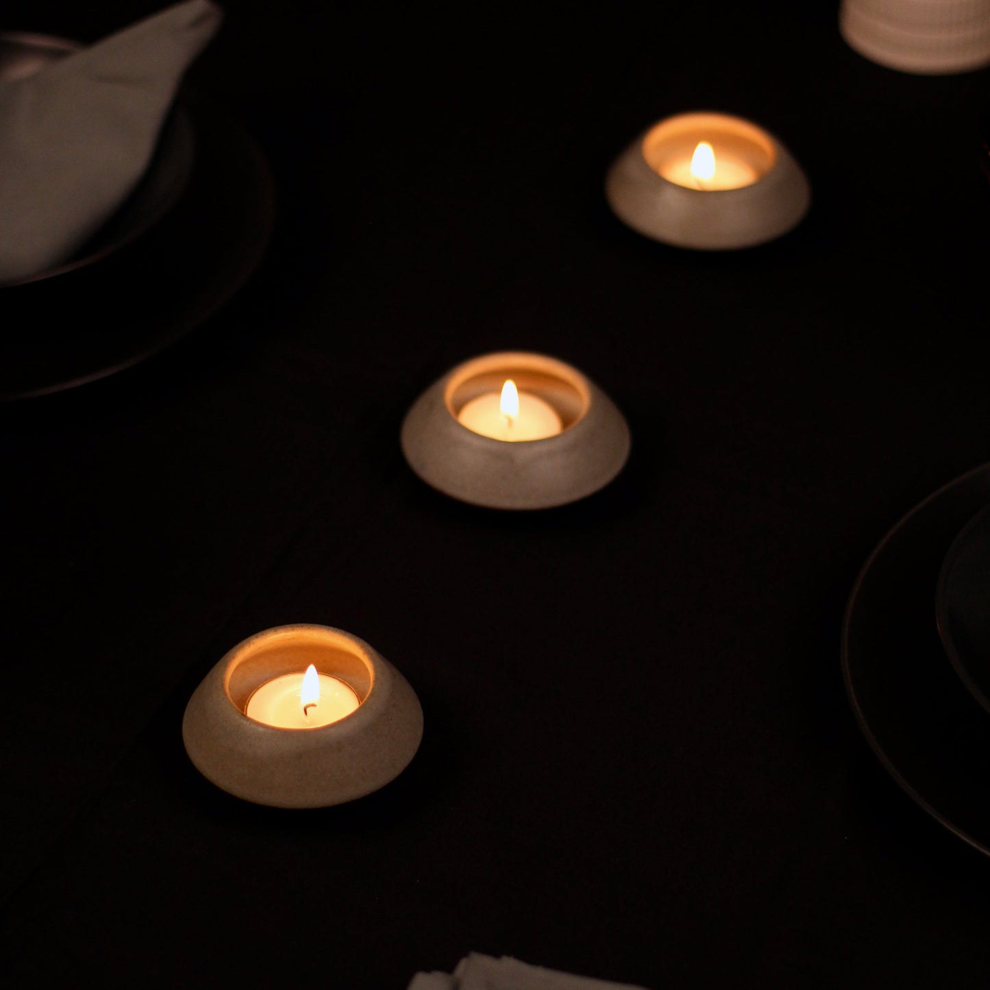 Set of three lit tealight candles on a dark surface with cutlery and plates.