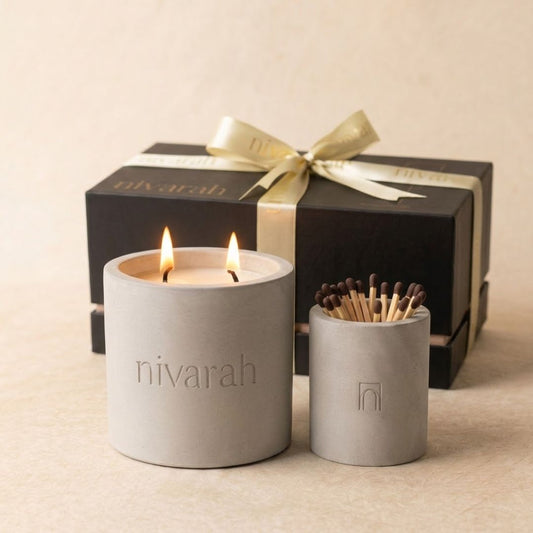 A Nivarah soy wax scented candle in a matte gray cylindrical jar with two lit wicks and a matchstick jar holder, with a black gift box and gold ribbon in the background.