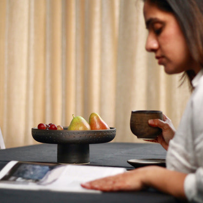 Person sitting at a table with a pedestal bowl in the background and a cup in hand, reading a magazine.