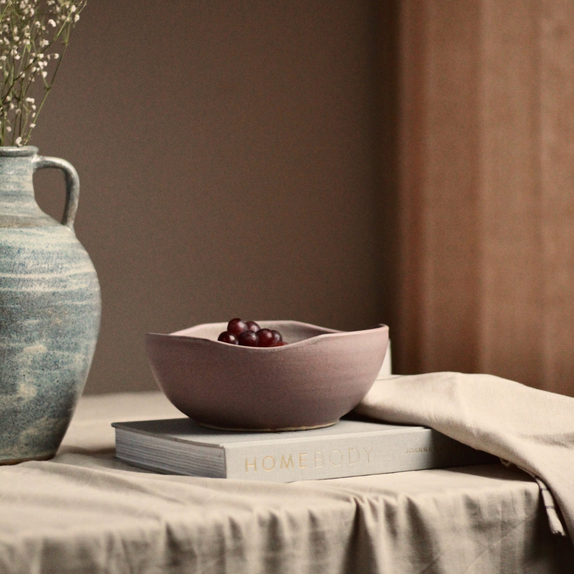 Ceramic bowl with grapes on a book next to a textured vase on a table with a brown background