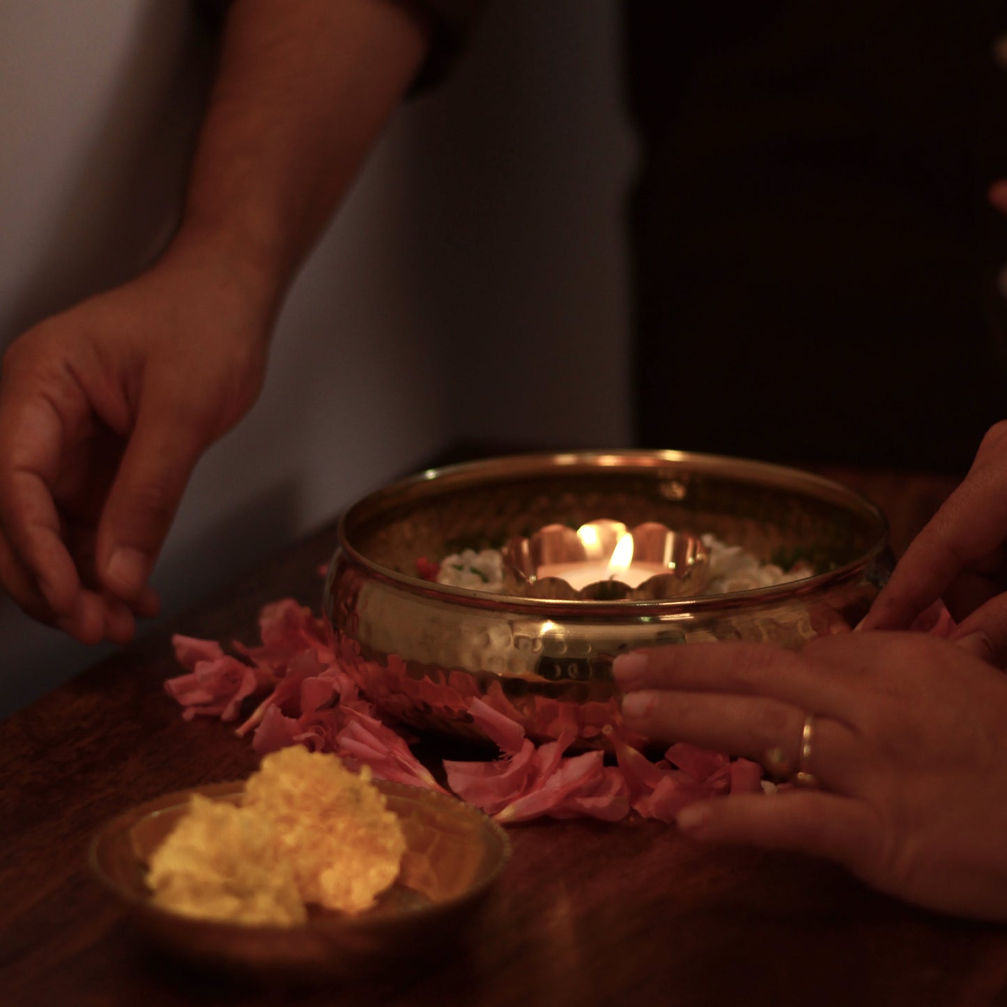 Hands interacting with a lit brass lamp and small brass plate dish on a wooden surface.