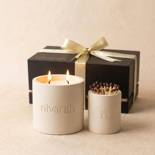 A Nivarah soy wax scented candle in a white cylindrical jar with two lit wicks and a matchstick jar holder, with a black gift box and gold ribbon in the background.