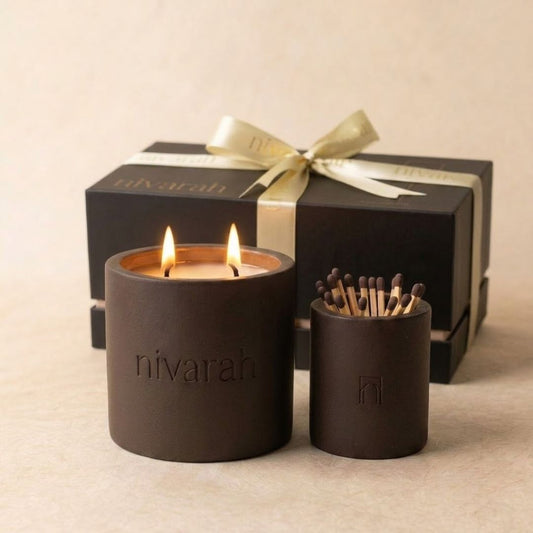 A Nivarah soy wax scented candle in a coffee brown cylindrical jar with two lit wicks and a matchstick jar holder, with a black gift box and gold ribbon in the background.
