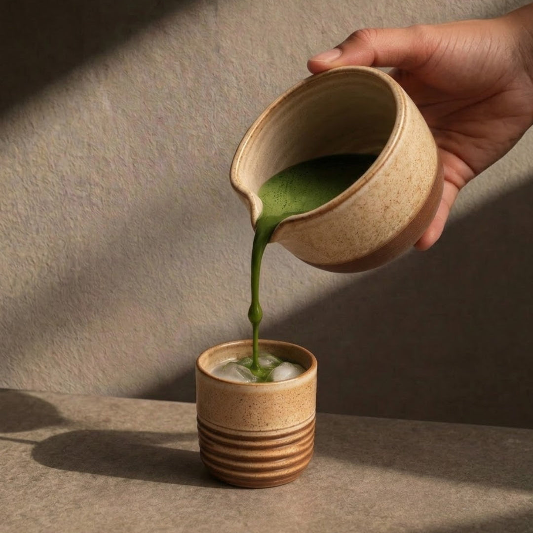 A hand pours green matcha tea from a beige ceramic pitcher into a small brown ceramic cup with horizontal ridges, set against a textured neutral background with dramatic shadows.