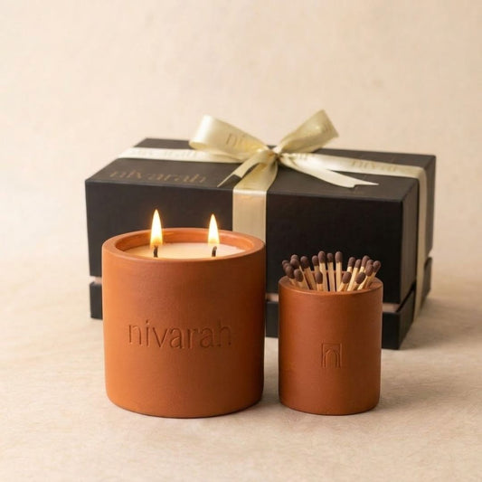 A Nivarah soy wax scented candle in a terracotta colored cylindrical jar with two lit wicks and a matchstick jar holder, with a black gift box and gold ribbon in the background.
